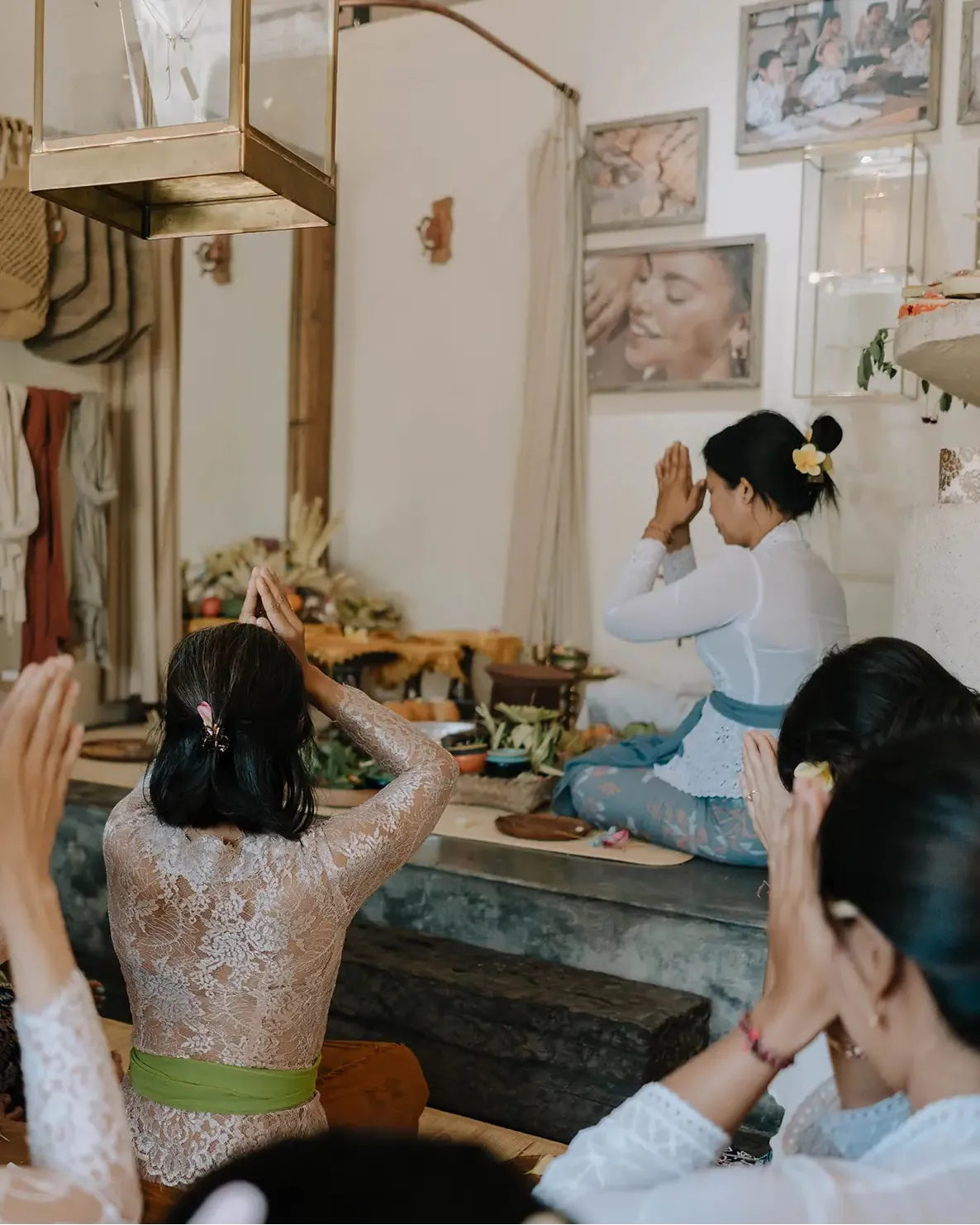 A group of women in traditional Balinese attire participate in a jewelry blessing ceremony, with one woman seated on a raised platform in the background and others standing with their hands raised in prayer. The setting features a warm, rustic interior with wooden furniture, framed wall art, and natural light.